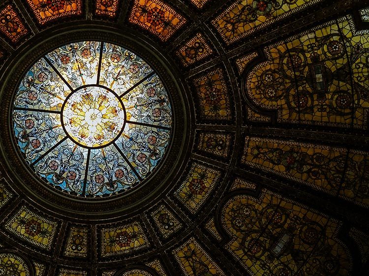 Looking up at the inside of a building's domed roof with intricate stained-glass panels