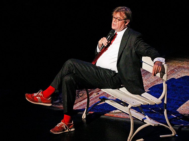 Garrison Keillor sitting on a bench wearing red trainers