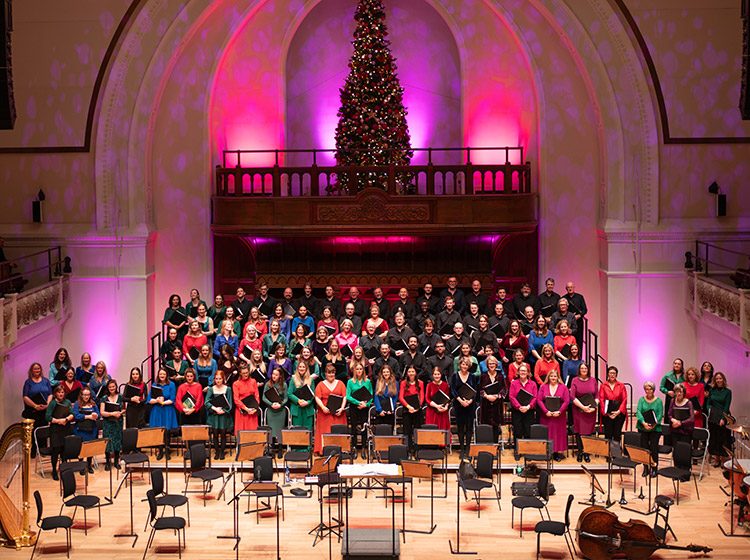 The City of London Choir on stage at Cadogan Hall with the women dressed in bright colours and the men in black