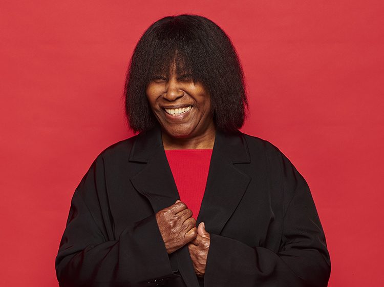A smiling Joan Armatrading dressed in a black jacket and stood against a red background