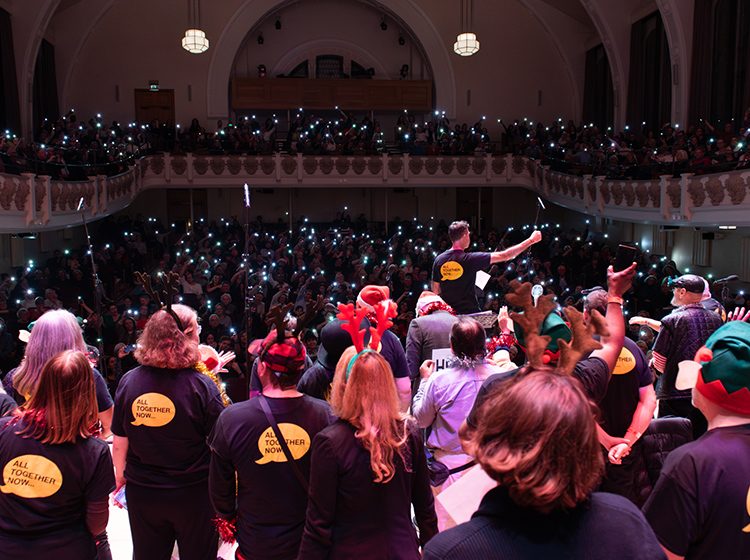 View from behind of The Choir with No Name on stage at Cadogan, and the audience behind holding up their mobile phone torches to create a glittering background