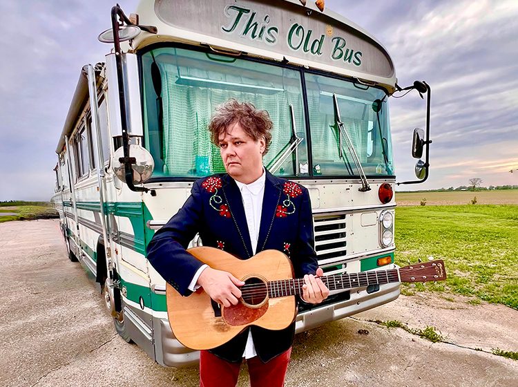 Ron Sexsmith holding his guitar, standing in front of 'This Old Bus'