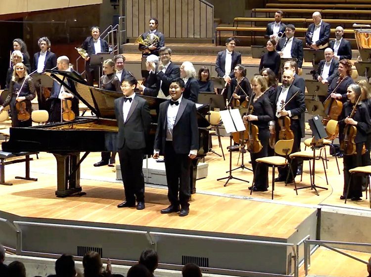 Carey Tan & Kevin Tan on stage with an orchestra behind them